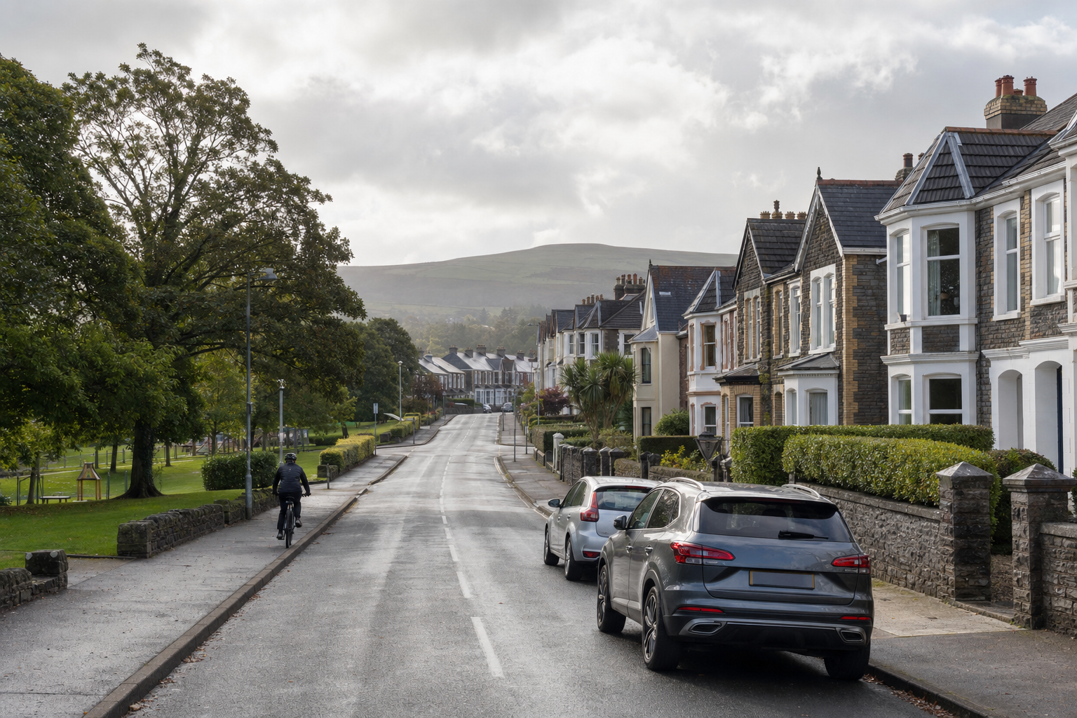 A cyclist rides down a residential street lined with parked cars and Victorian houses, with hills in the background.