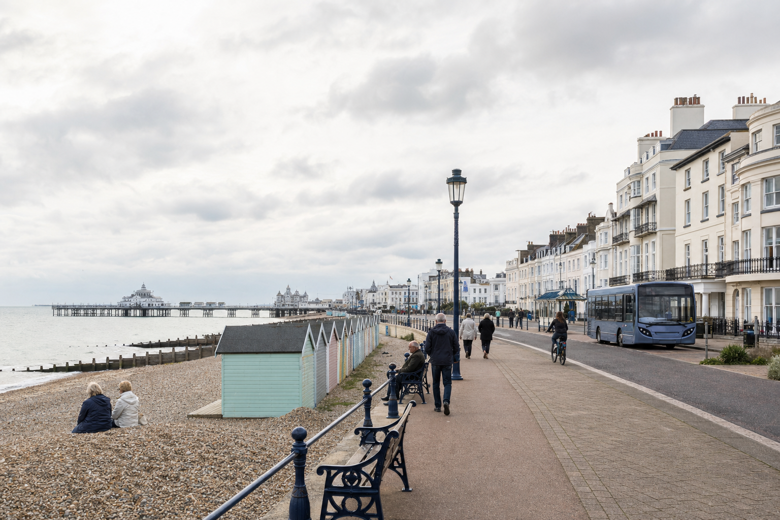 Seaside promenade with people walking, colourful beach huts, and a distant pier under a cloudy sky.