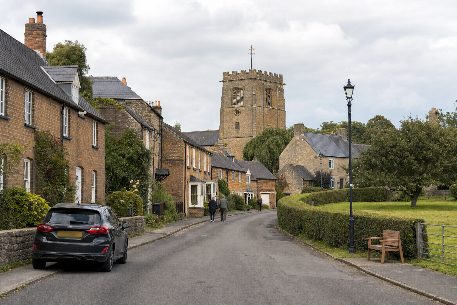 Quaint village street with a stone church tower in the distance and a parked car on the left.