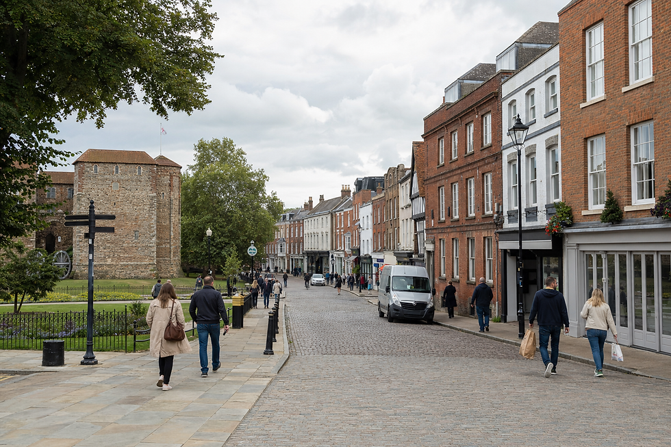 People walk along a cobbled street lined with historic buildings and a round castle tower in the background.