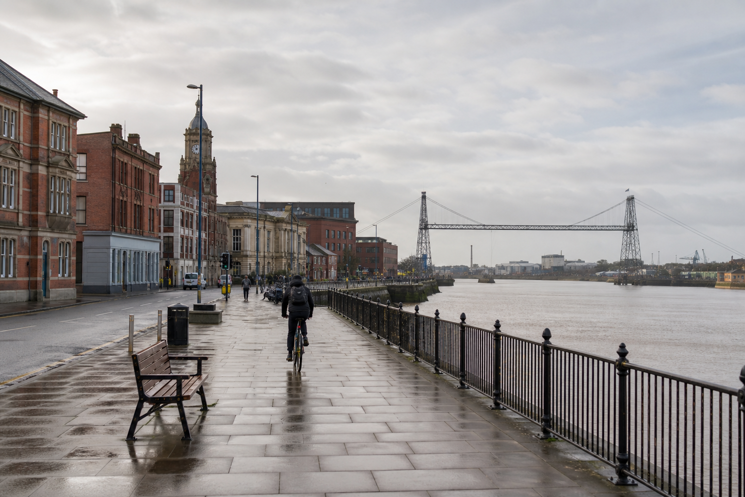 A cyclist rides along a wet riverside path with Victorian architecture and a transporter bridge in the background.
