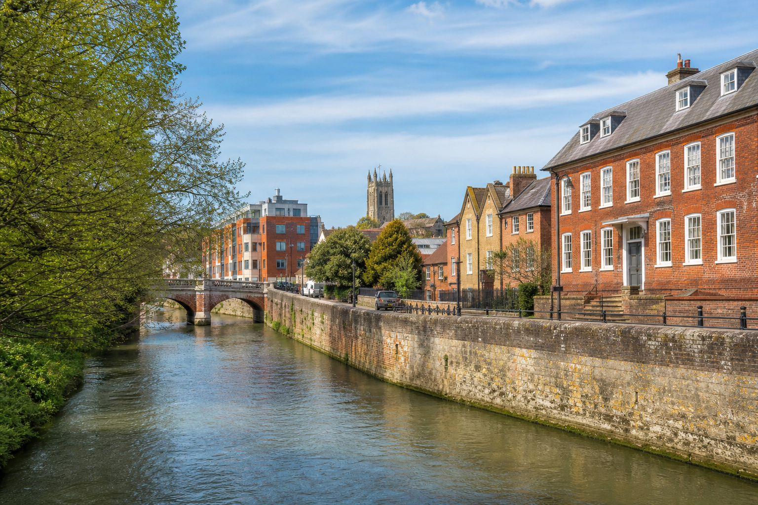 River scene with brick buildings, a stone bridge, and a distant church tower under a blue sky.