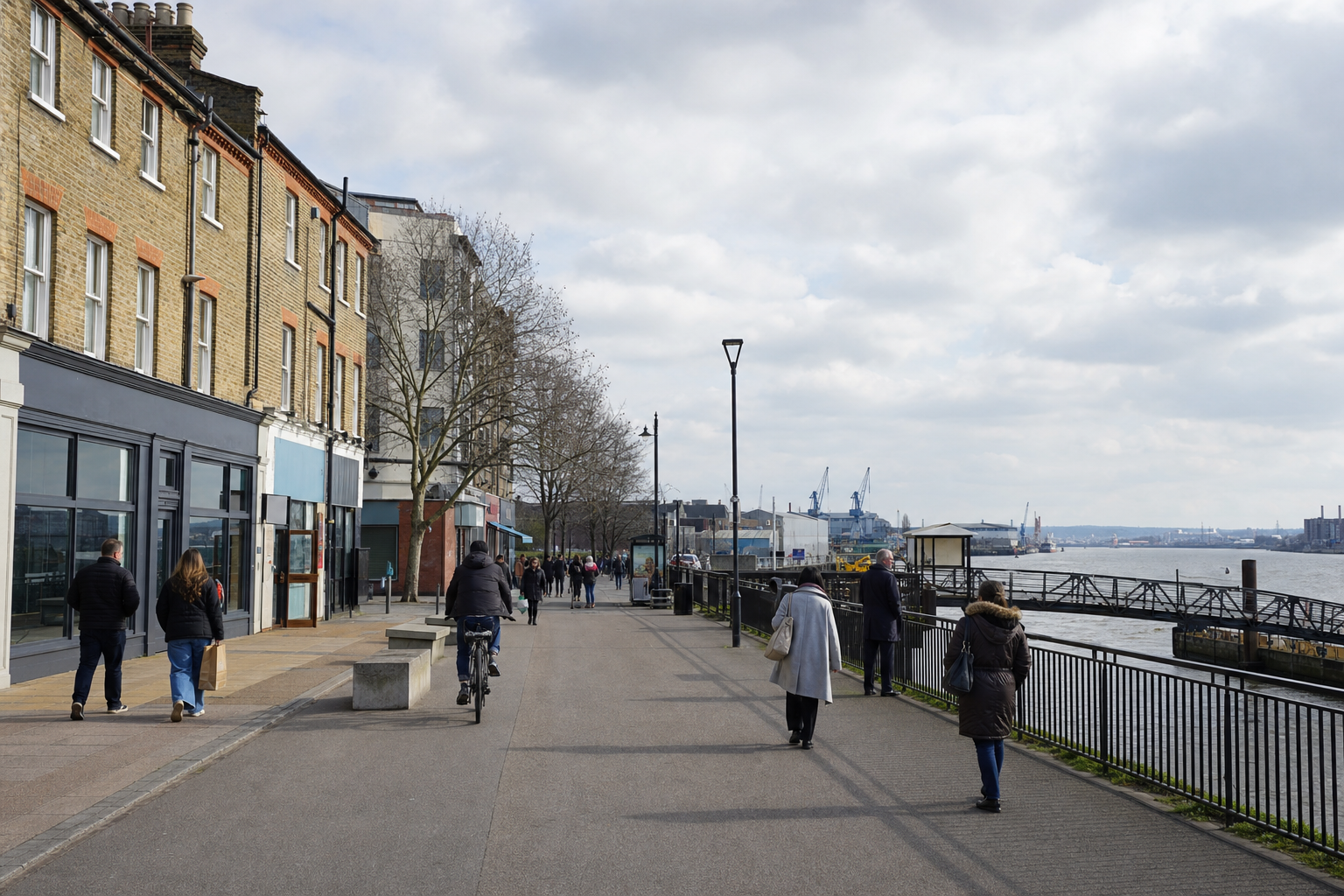 People walking and cycling along a riverside promenade with buildings and industrial cranes in the background.
