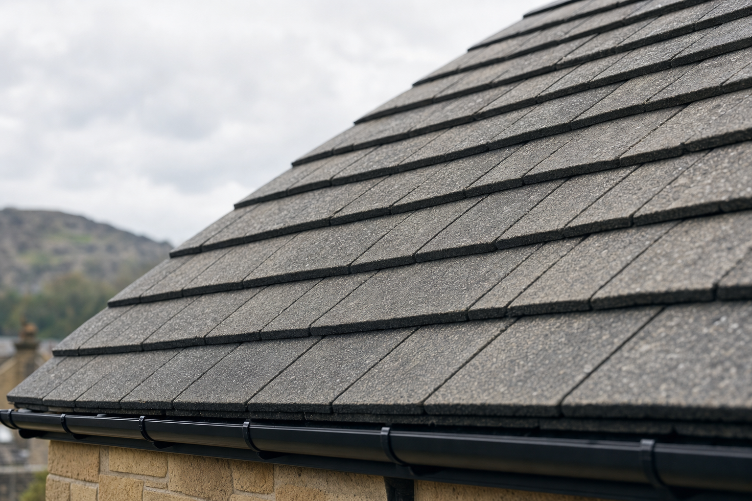 A close-up of a grey tiled roof with a black gutter against a cloudy sky backdrop.