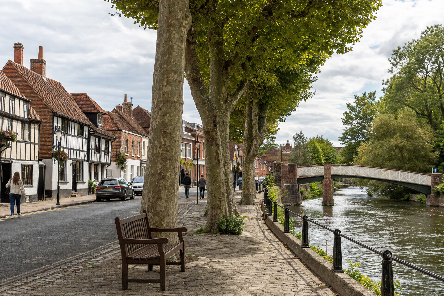 Historic riverside street with half-timbered houses, tree-lined promenade, and an arched footbridge under a cloudy sky.