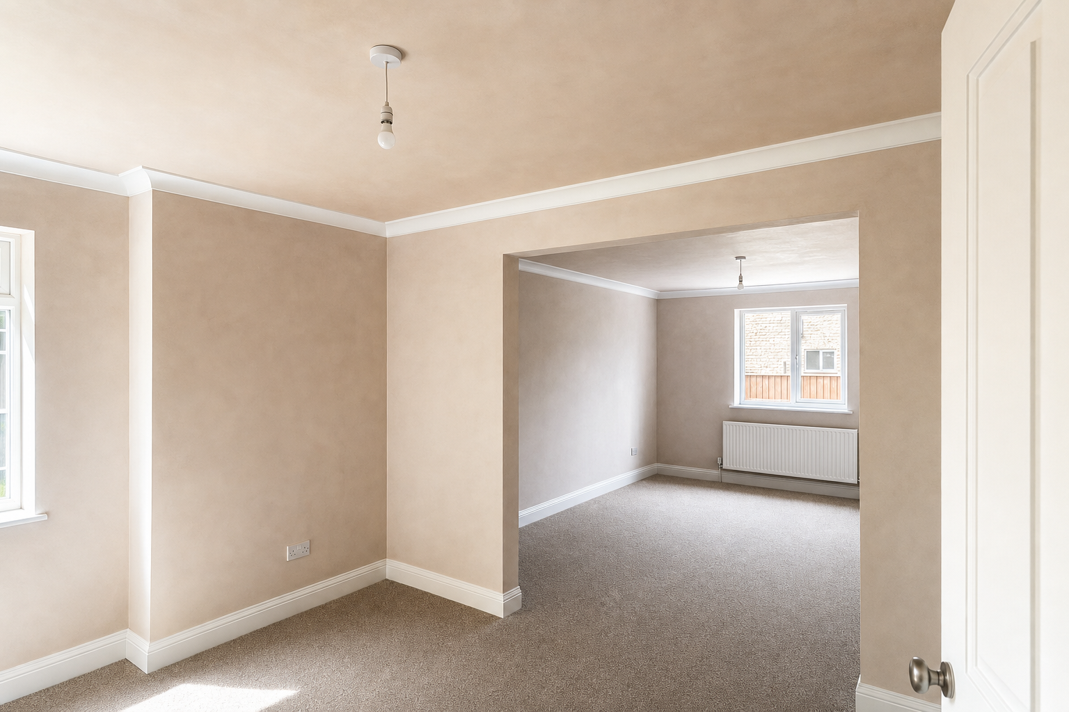 Empty beige room with carpet and large window, leading to a smaller adjacent room with radiator.