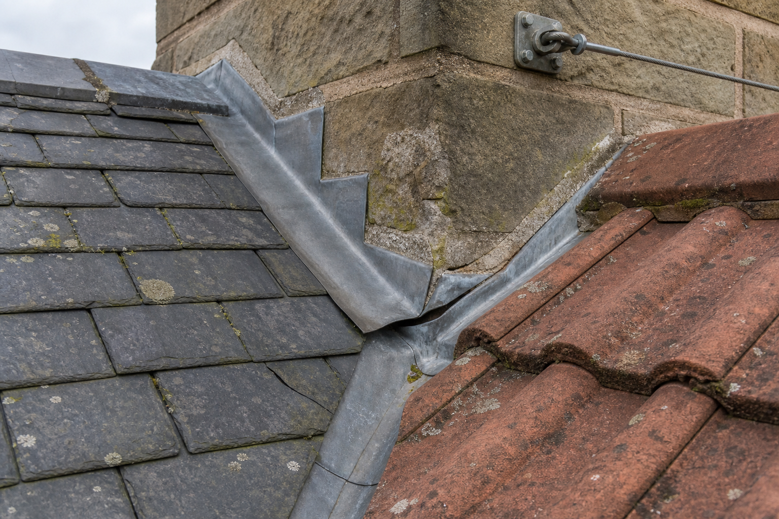 Slate and terracotta tiled roofs intersect at a stone chimney with lead flashing for waterproofing.