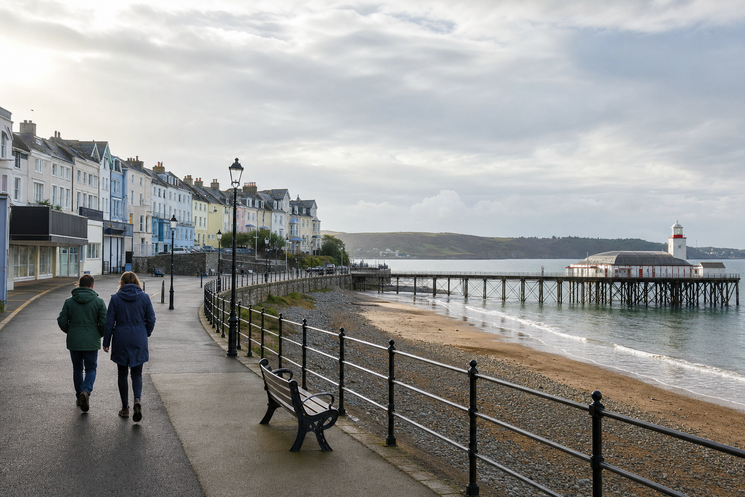 Seaside promenade with two people walking, colourful buildings, and a pier extending over the calm sea.