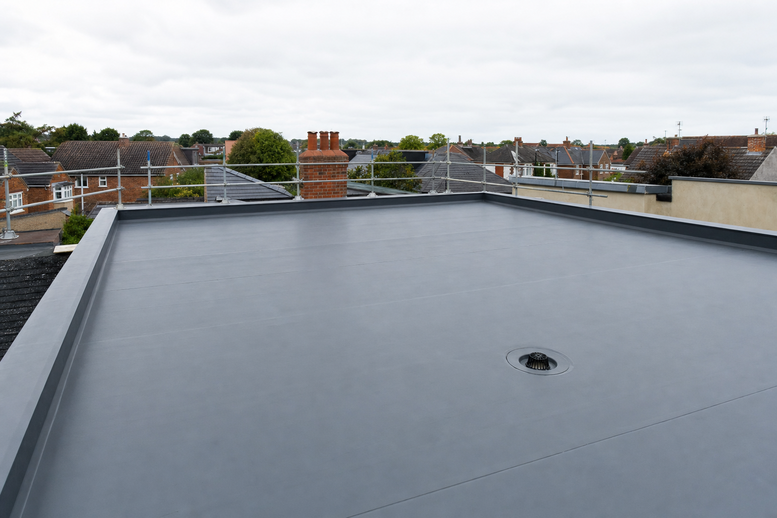 Flat rooftop with a grey water drainage system, surrounded by railings, overlooking a residential area.