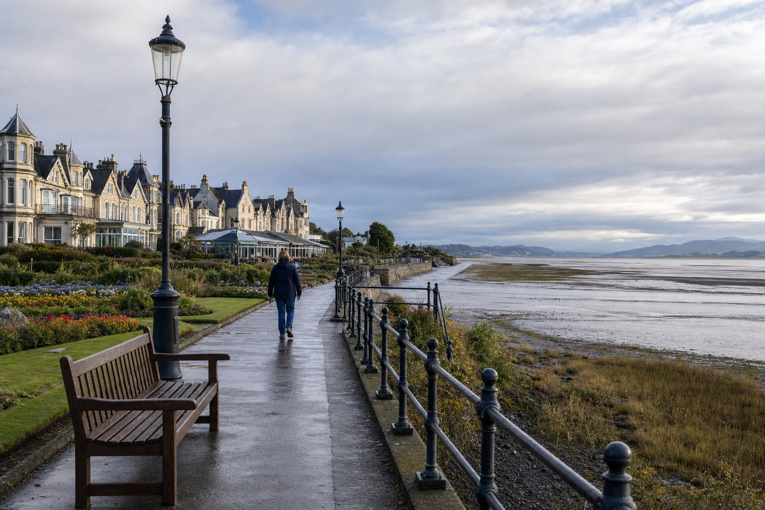 A person walks along a promenade by the sea, with Victorian-style houses and a bench under a cloudy sky.