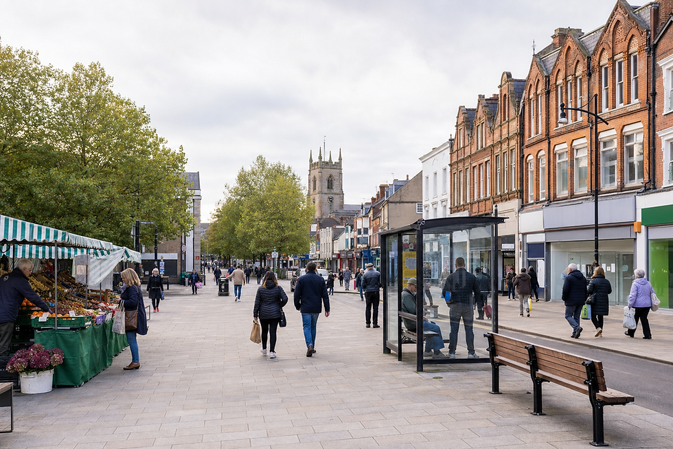 A busy pedestrian street features market stalls, people walking, and a historic church tower in the background.