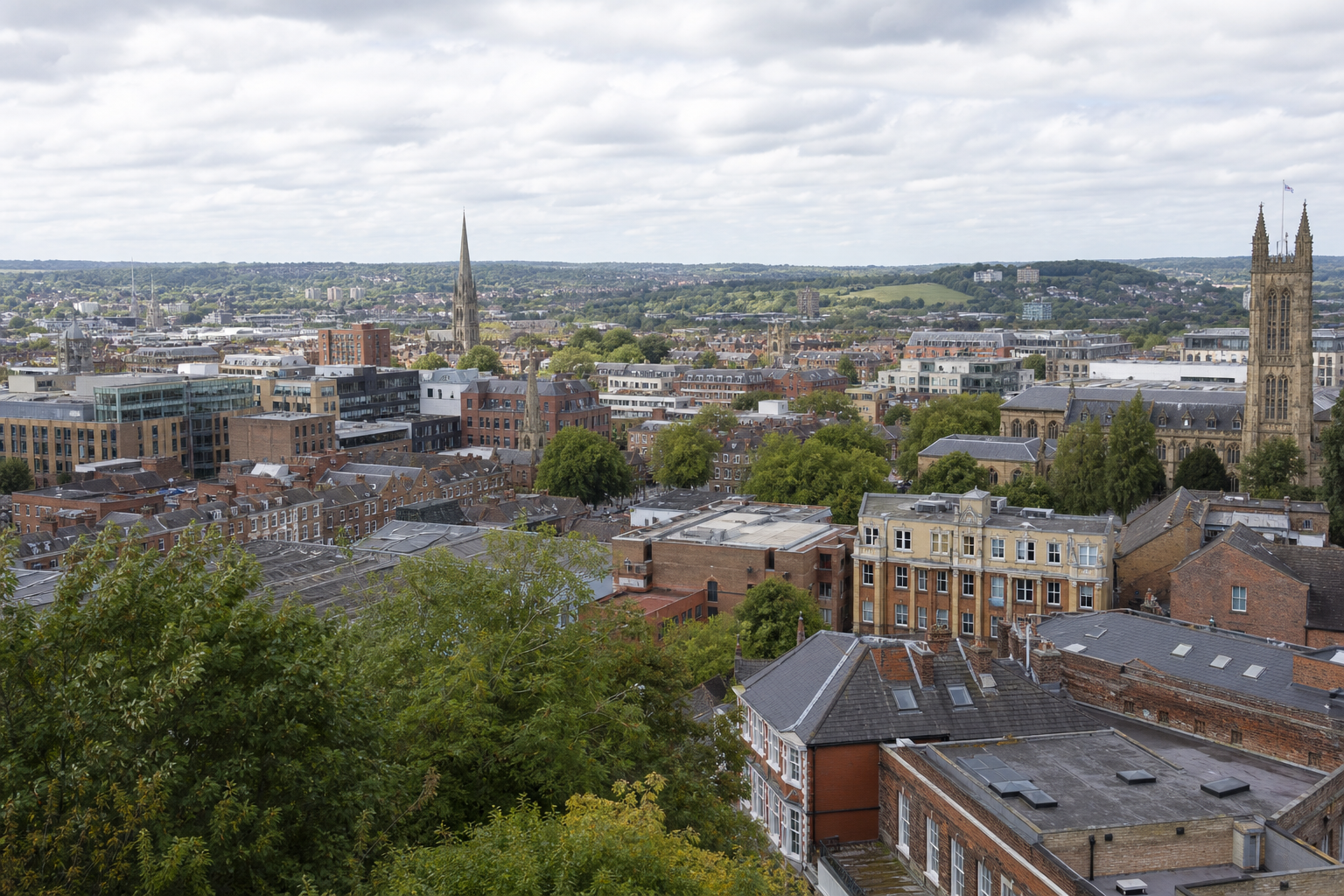 Aerial view of a cityscape with historical buildings, trees, and distant rolling hills under a cloudy sky.