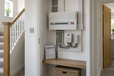 A modern electrical consumer unit with circuit breakers is mounted on a wall in a bright hallway.