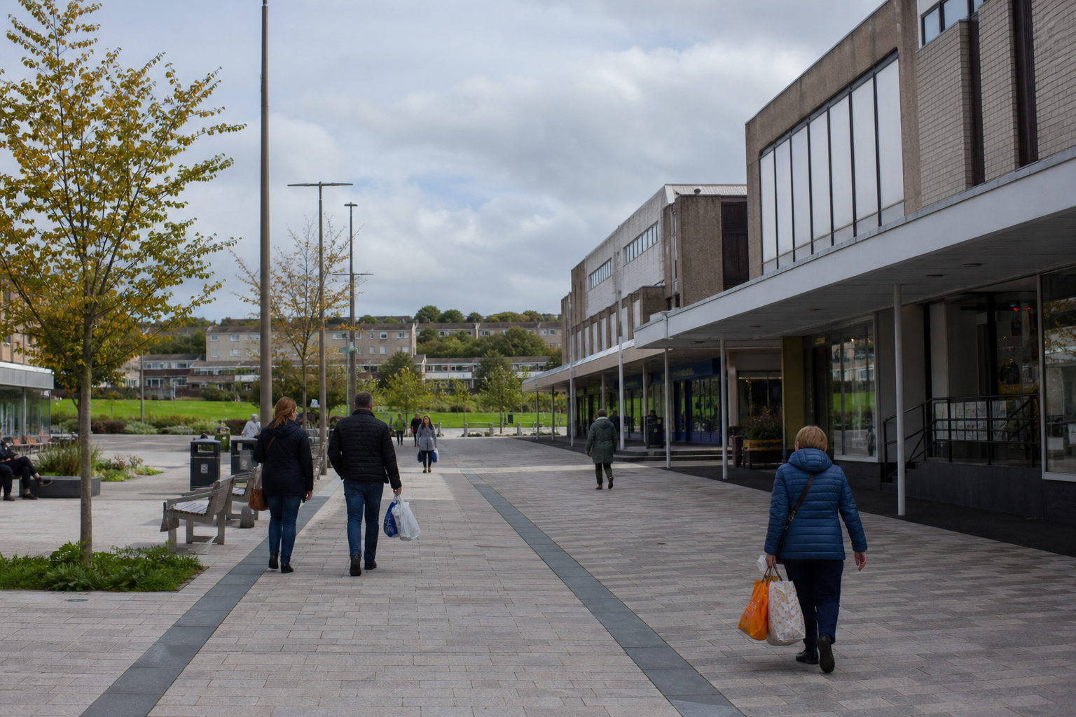 Pedestrians walk through an outdoor shopping area with trees and benches on a cloudy day.