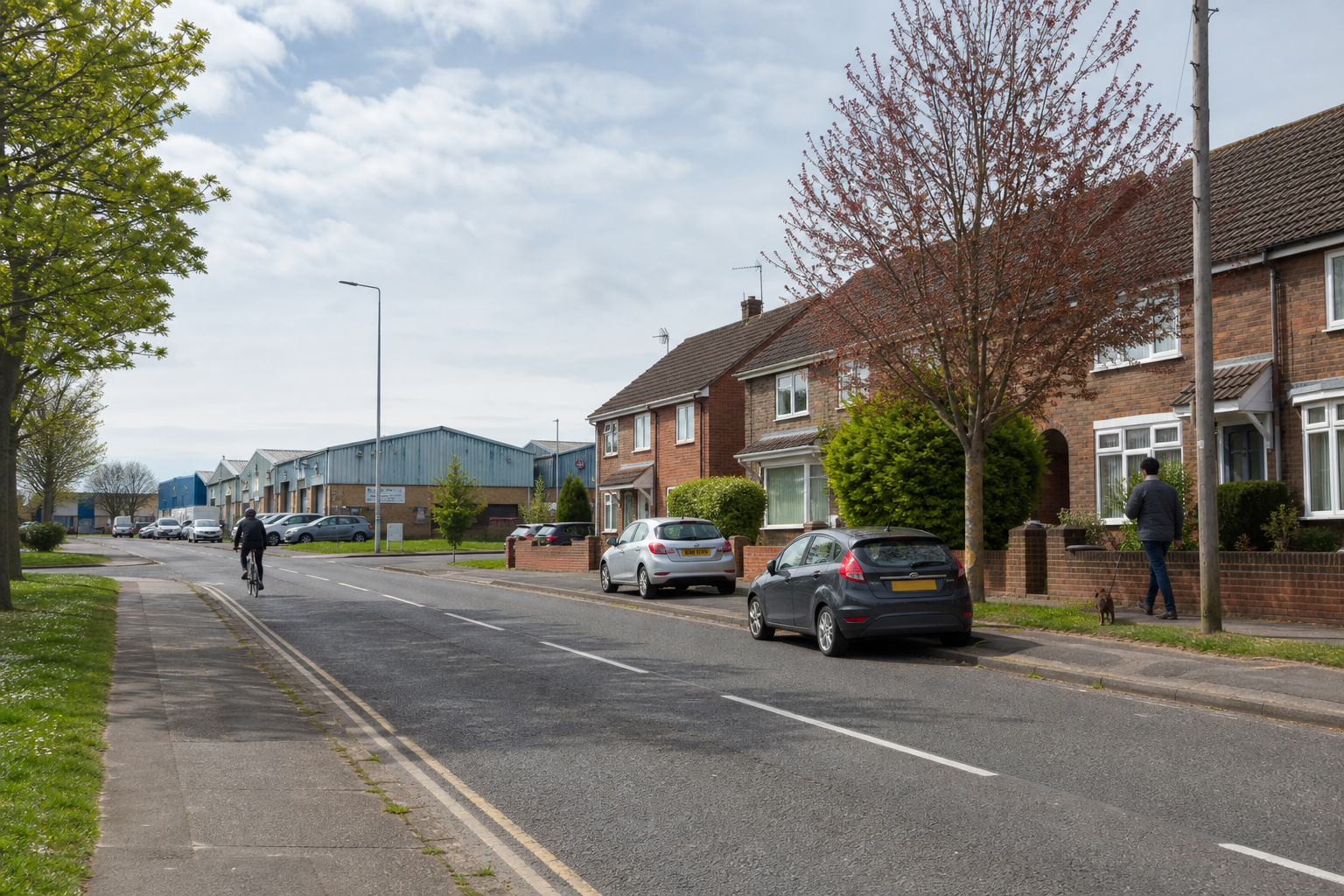 A cyclist rides along a suburban street with parked cars and industrial buildings in the background.
