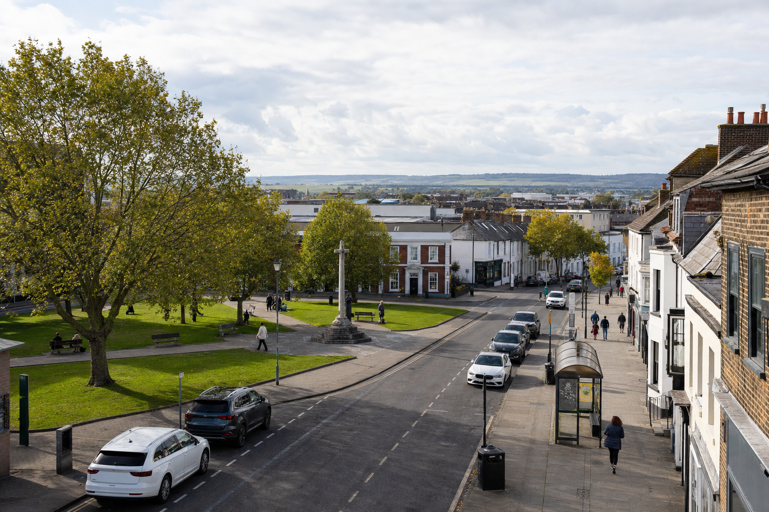 A quiet street scene with cars parked along the kerb, people walking, and a small green park with a memorial in the centre.