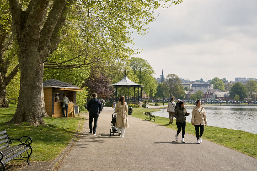 People walking along a path by a lake in a park, with a bandstand and town buildings in the background.