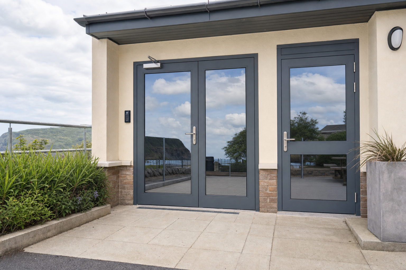 Modern building entrance with a glass door reflecting a coastal landscape, surrounded by greenery.
