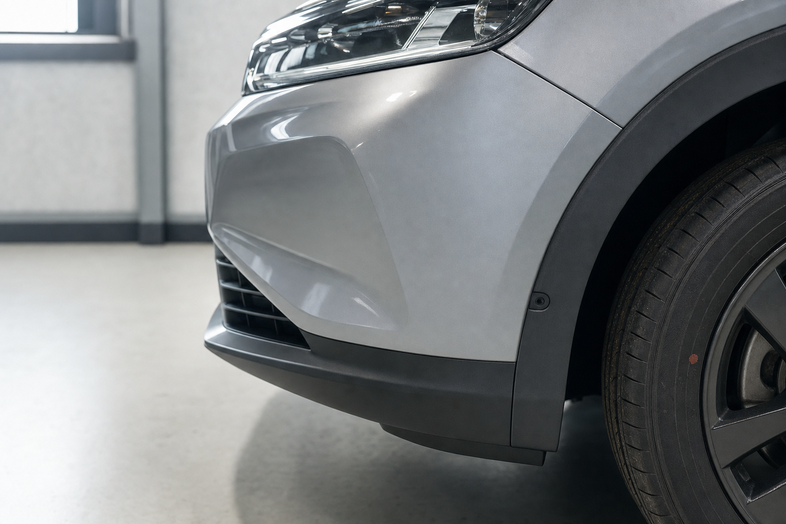 Close-up of a silver car's front left bumper, headlight, and tyre in a garage setting.