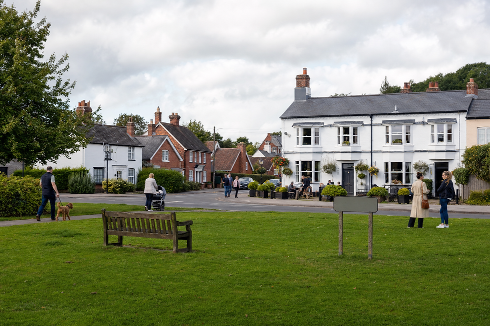 Village green with people walking and chatting near charming red-brick and whitewashed houses on a cloudy day.