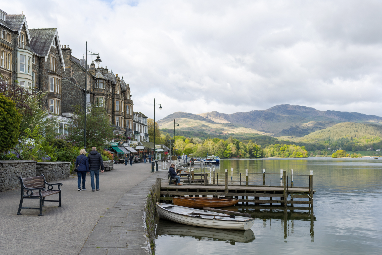 Couple walking along a scenic lakeside promenade with mountain views and boats moored by the pier.