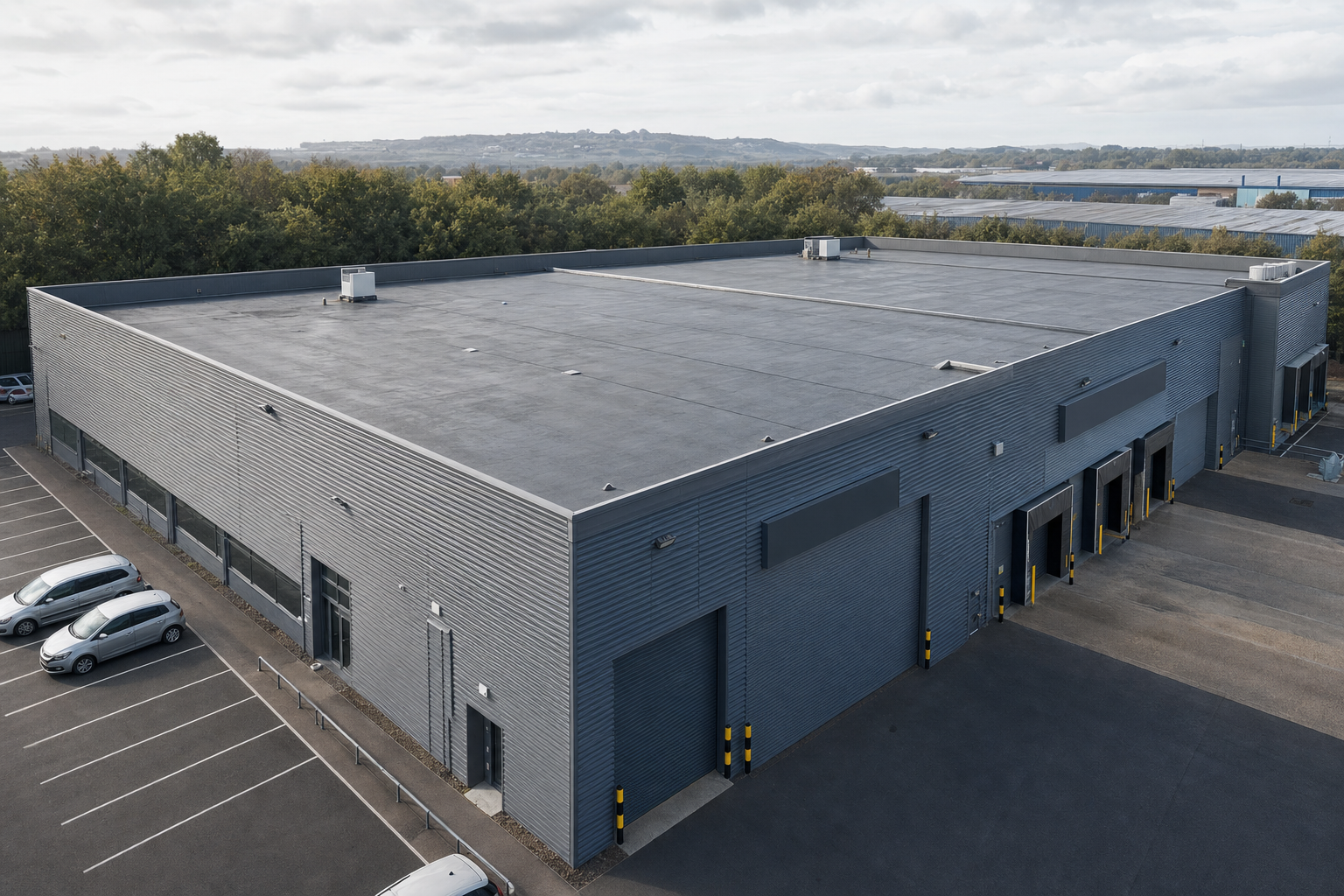 Large industrial warehouse with a grey exterior, adjacent parking spaces, and surrounded by trees.