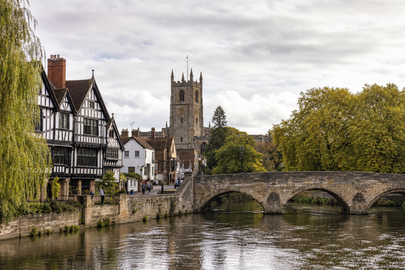 Historic stone bridge over a river with half-timbered houses and a church in the background, surrounded by autumn trees.