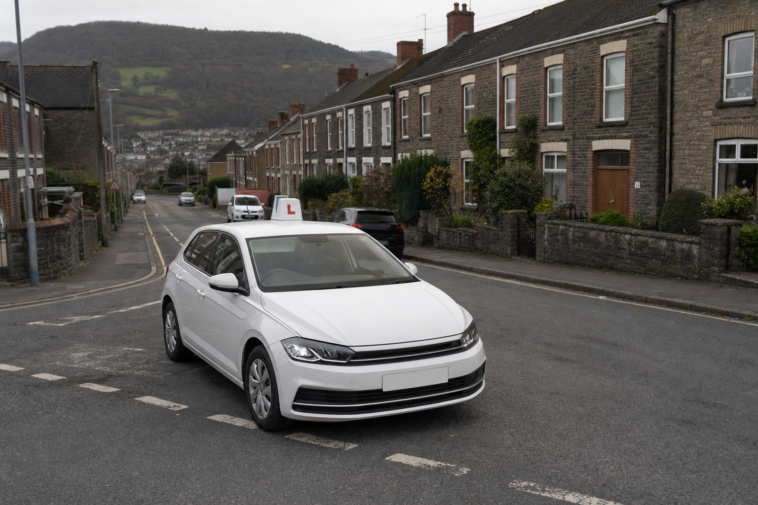 A white car with a learner driver sign on the roof is parked on a residential street with terraced houses and a hill in the background.