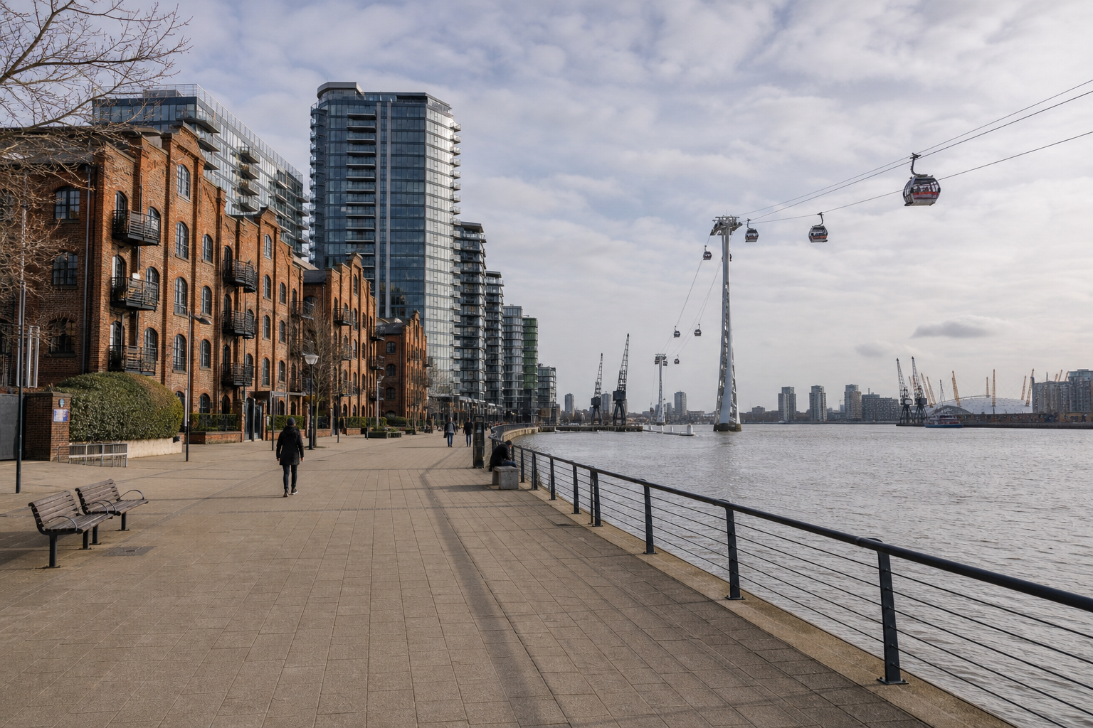 Riverfront pavement with people walking, flanked by brick and glass buildings, cable cars overhead, and cranes across the water.