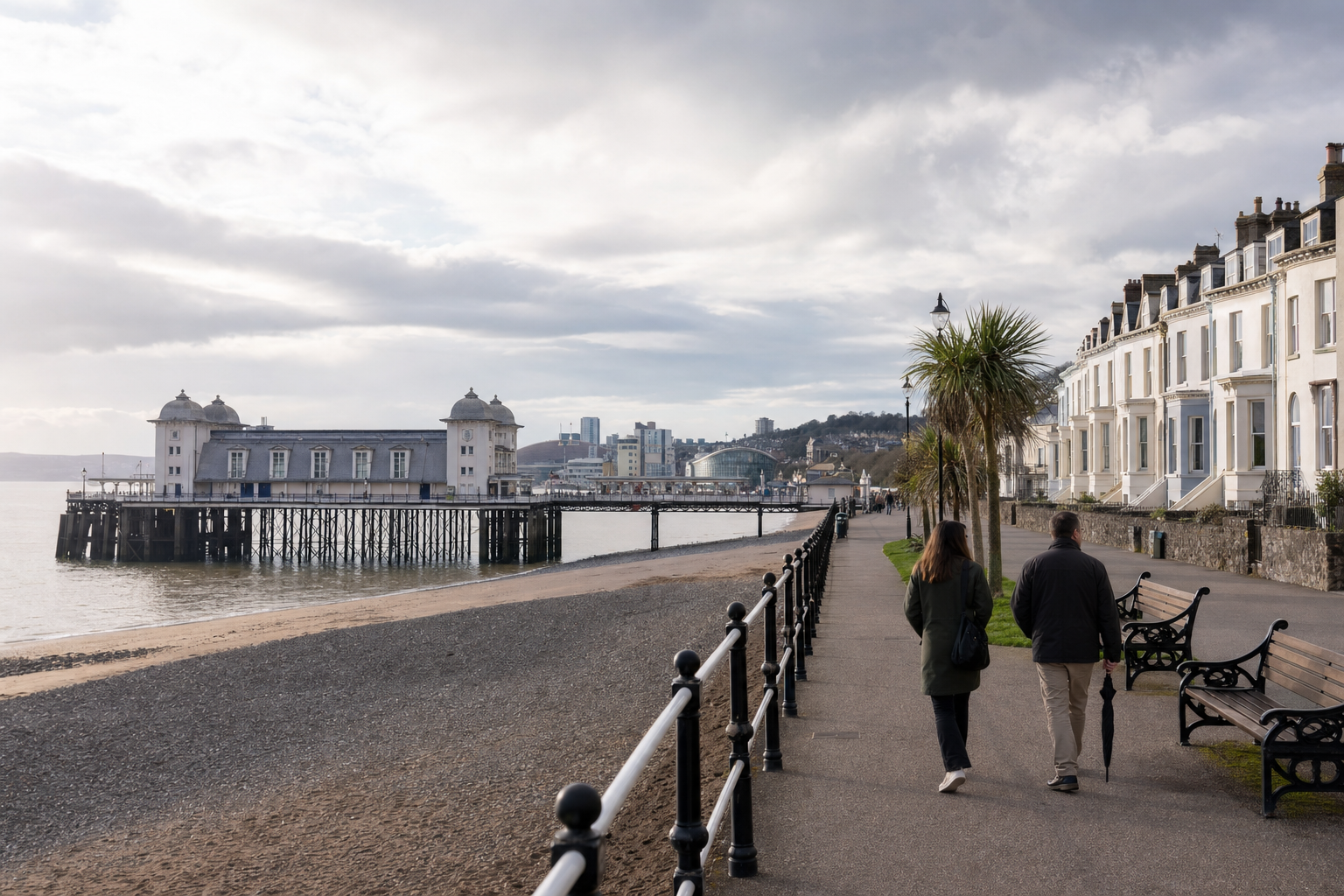 Two people stroll along a seafront promenade with a pier extending into the water and white terraced houses nearby.