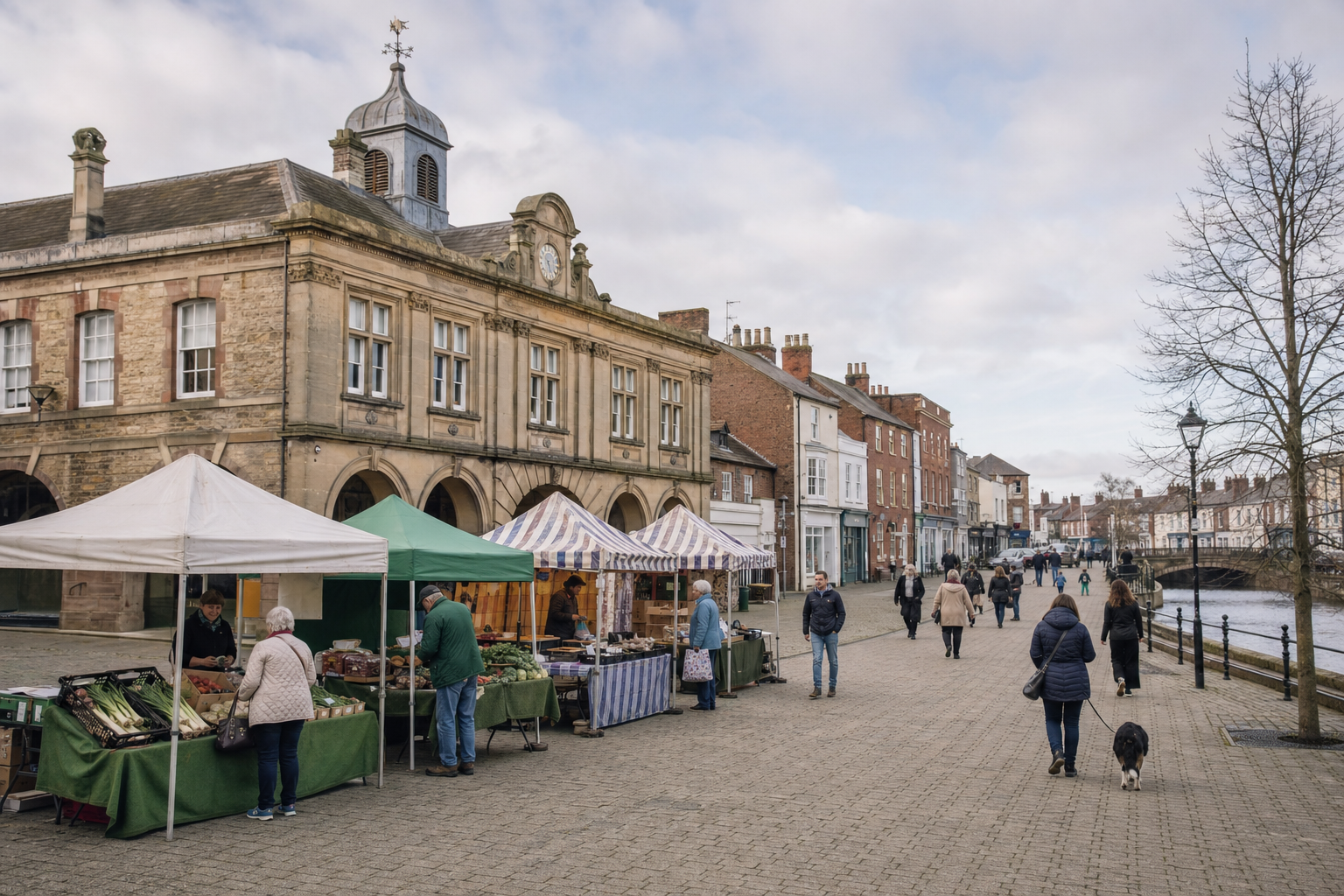 A street market with stalls and shoppers on a cobbled walkway beside historic buildings and a river.