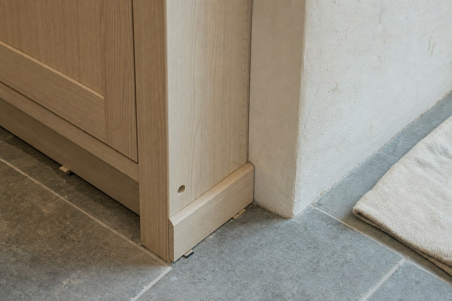 Close-up of wooden cabinet corner meeting a tiled floor and plastered wall with a beige rug nearby.
