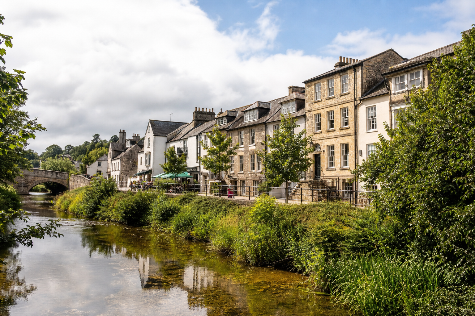 Quaint row of historic buildings alongside a serene river and stone bridge under a partly cloudy sky.