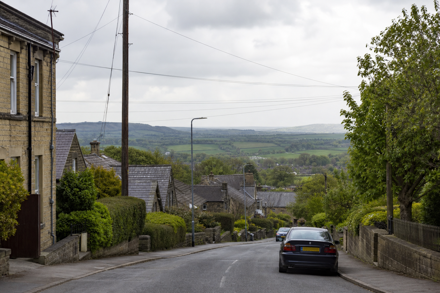 Countryside road with stone houses, parked cars, and rolling green hills in the background.