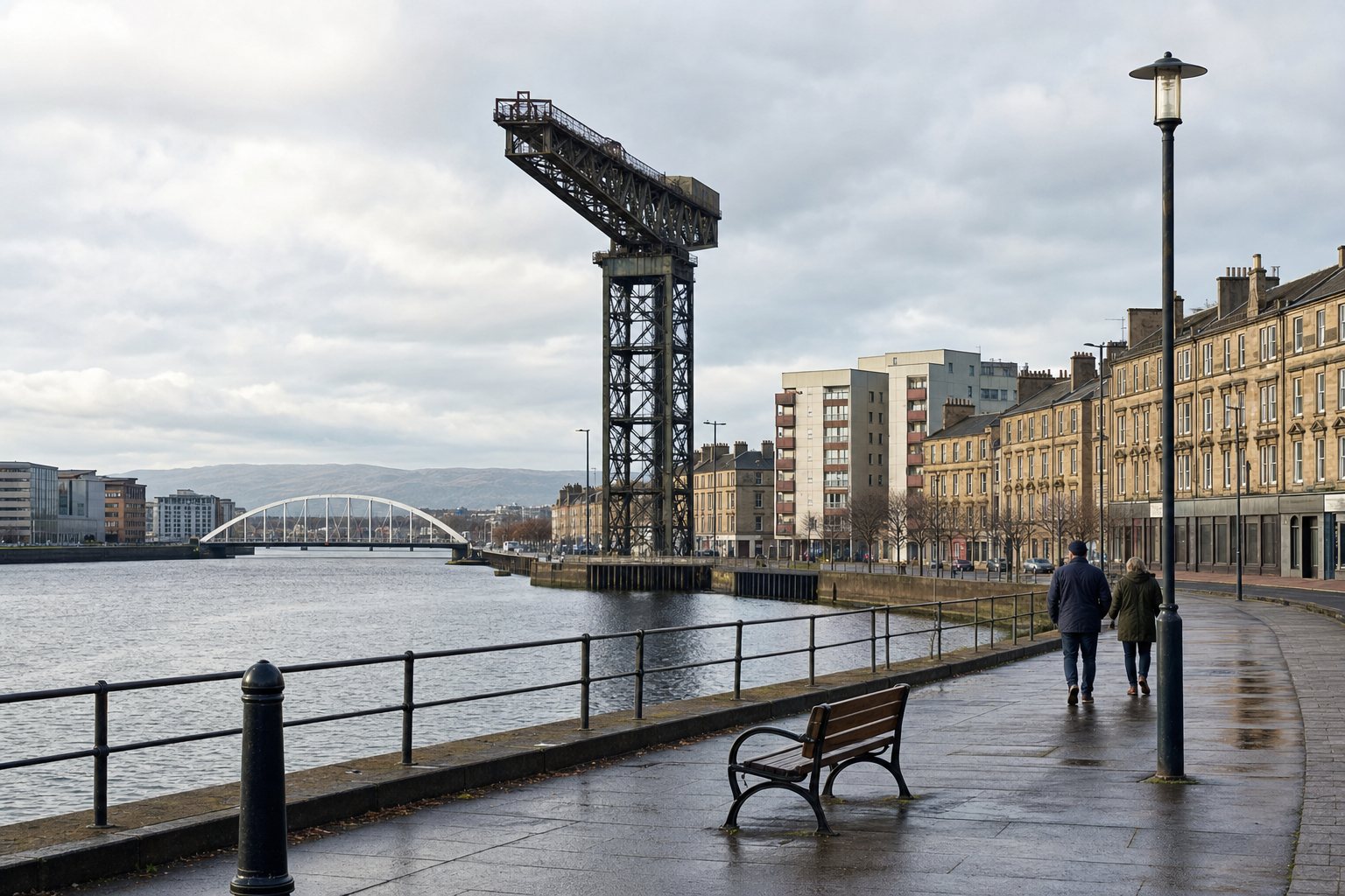 Two people walk along a riverside promenade beside a historic crane and buildings under a cloudy sky.