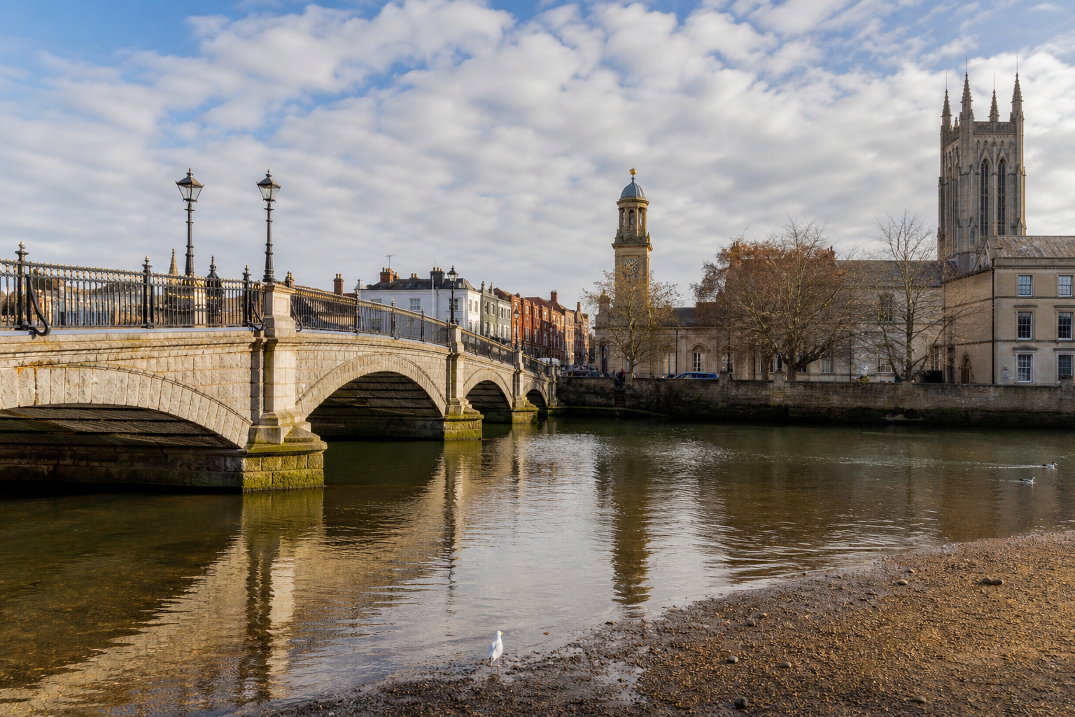 Stone bridge over a river, with historic buildings and a church tower in the background, under a cloudy sky.