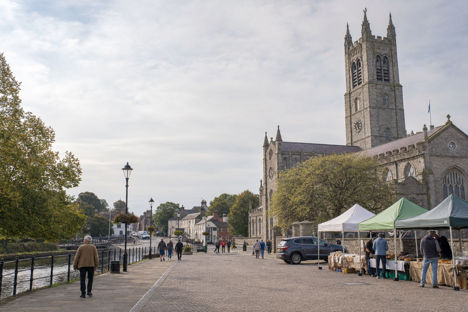 Street market with stalls in front of a historic stone church, people walking along the riverside path.