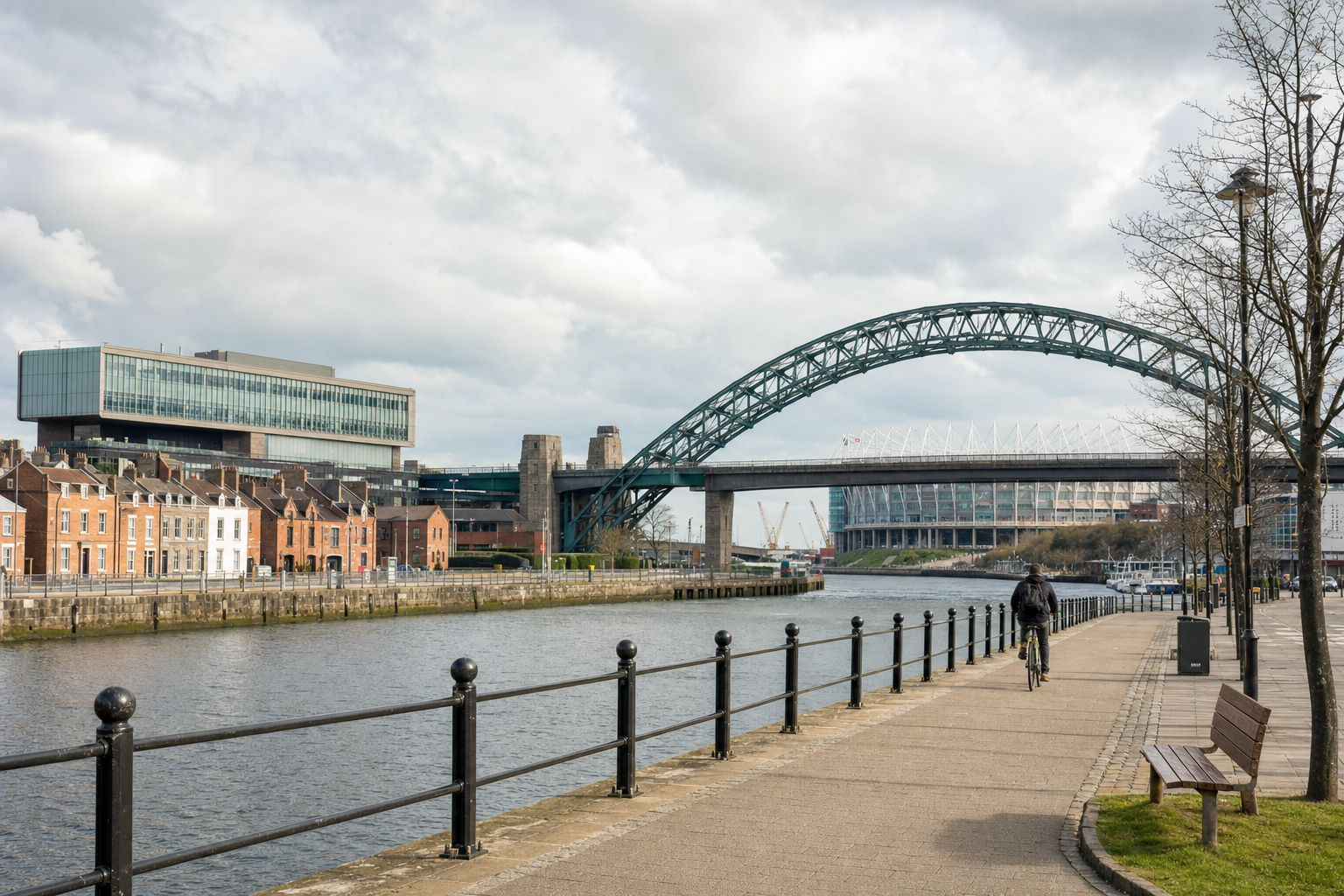 A cyclist rides along a riverside path with a bridge and stadium in the background.
