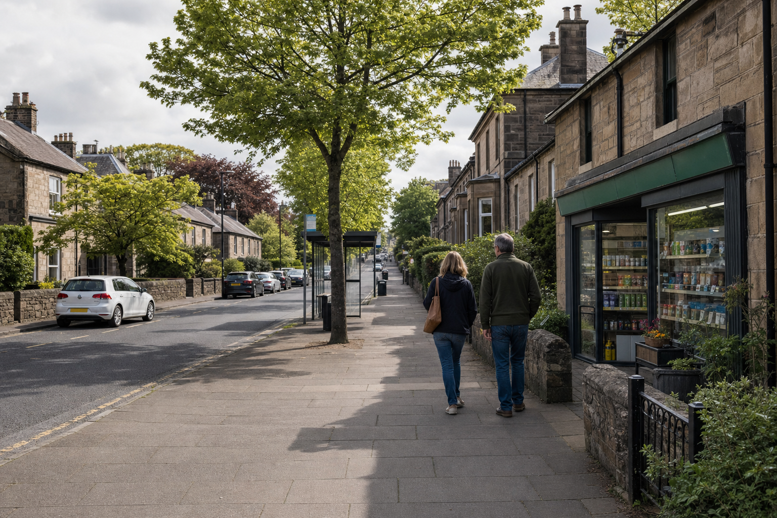 A couple walks down a tree-lined street with a row of stone houses and a small shop on a cloudy day.