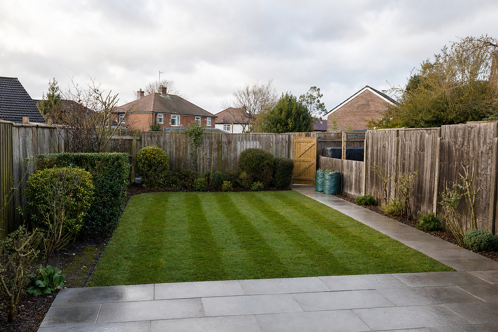 A neatly landscaped garden features striped grass, a wooden fence, and paving stones under a cloudy sky.