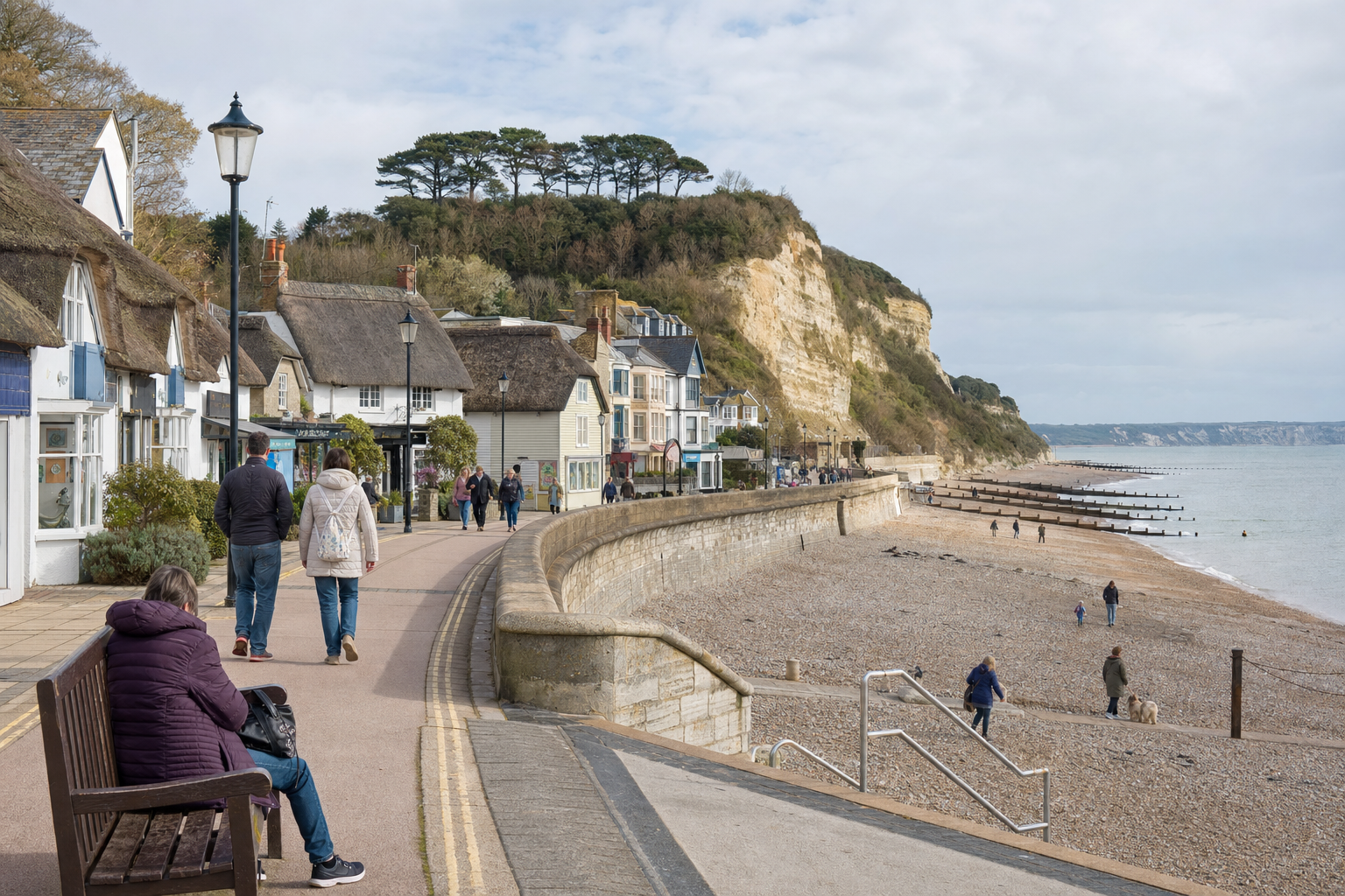 A coastal path lined with thatched cottages overlooks a pebble beach and distant cliffs, with people strolling and enjoying the view.