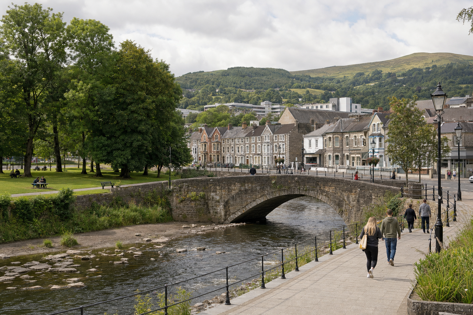 A stone bridge spans a river in a quaint town, with people walking along the riverside path and lush green hills in the background.