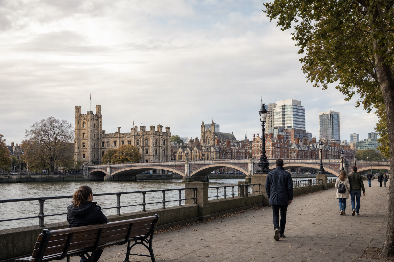 People walk along a riverside path with historic buildings and a bridge in the background on a cloudy day.