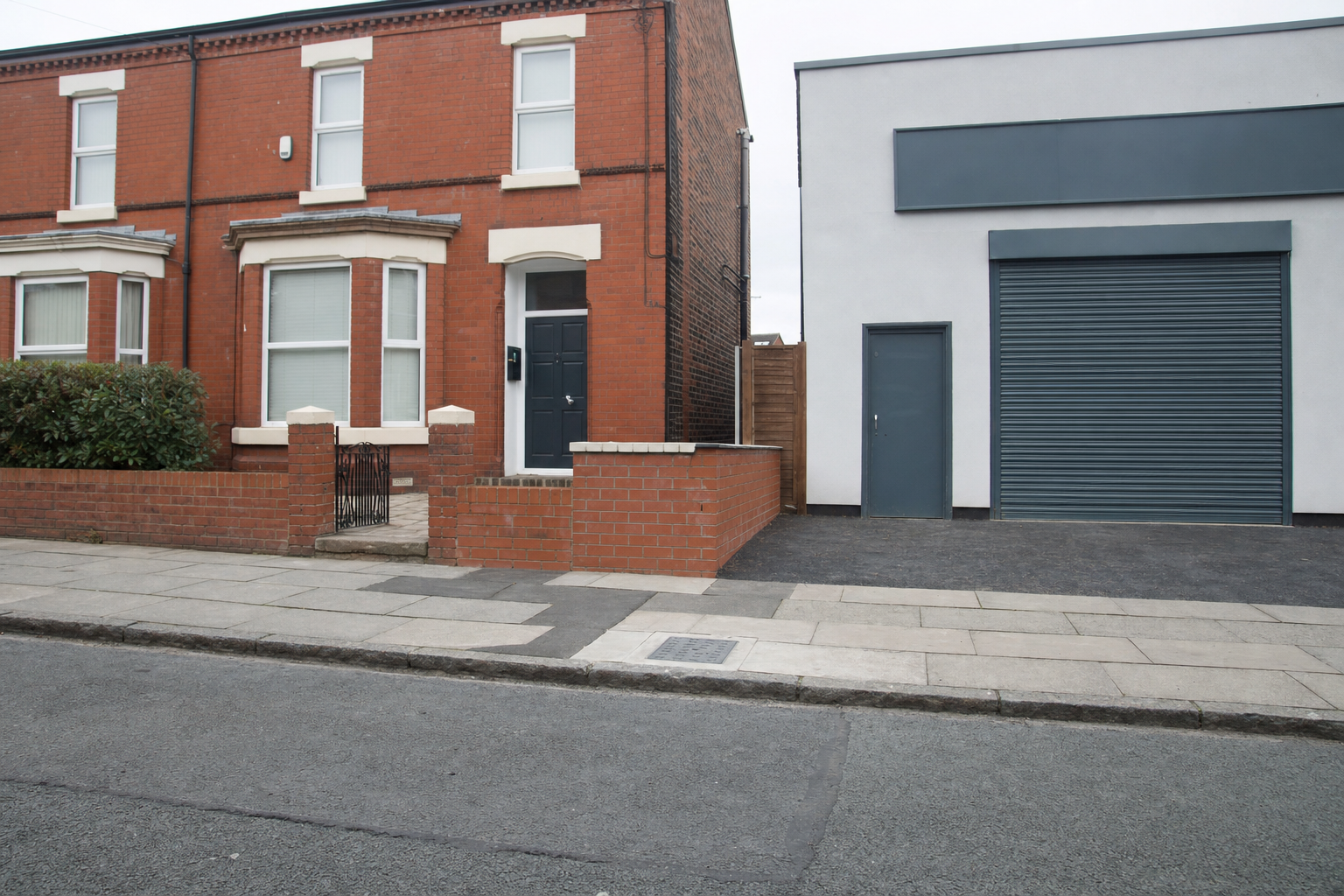 Red brick terraced house with a bay window next to a modern white building with a grey garage door.
