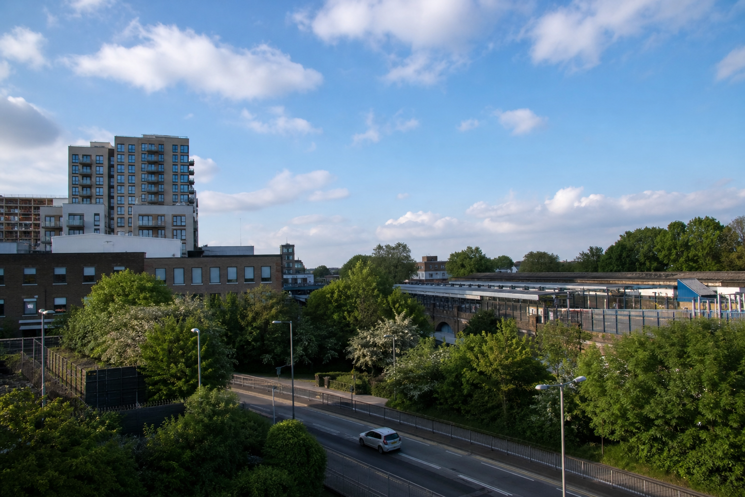 Urban landscape with a mix of buildings, trees, and a road under a blue sky with clouds.