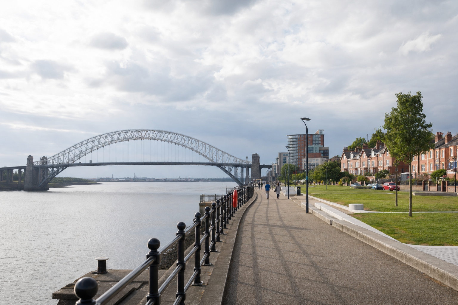 A riverside path with people walking, near a large arched bridge and residential buildings under a cloudy sky.