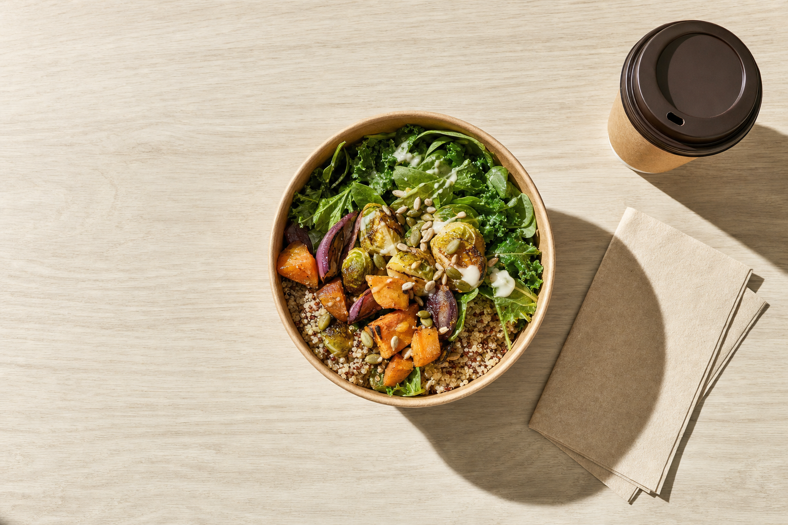 A wooden table hosts a quinoa and roasted vegetable salad beside a takeaway coffee cup and napkins.