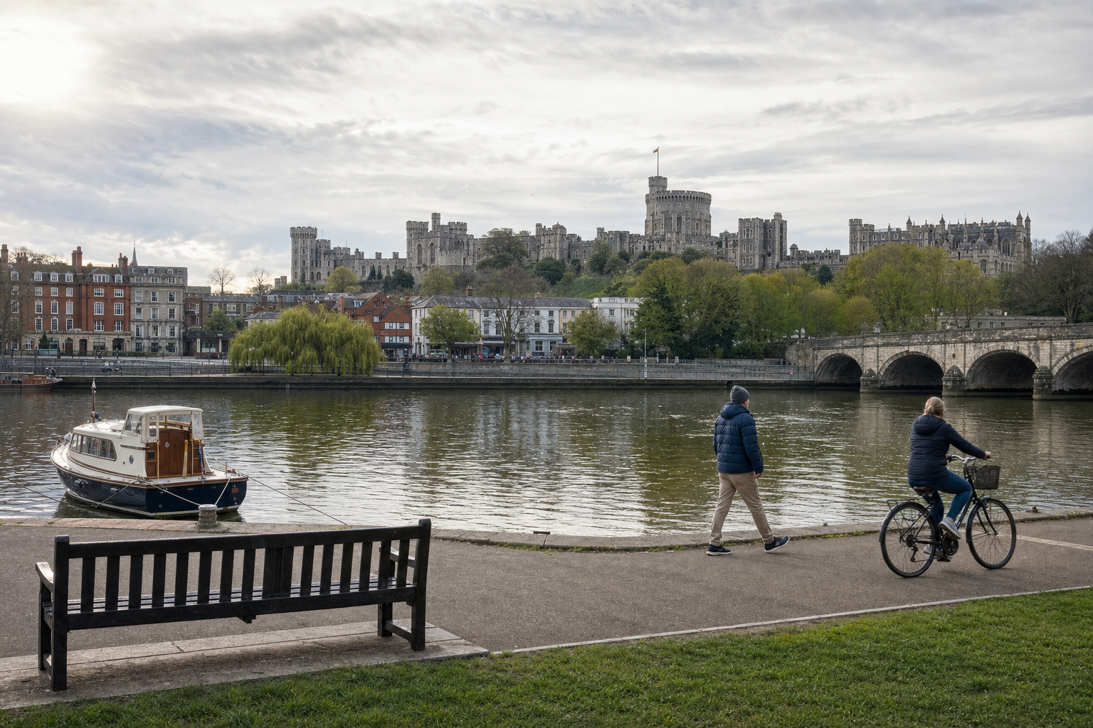 People walking and cycling by the river with Windsor Castle in the background under a cloudy sky.