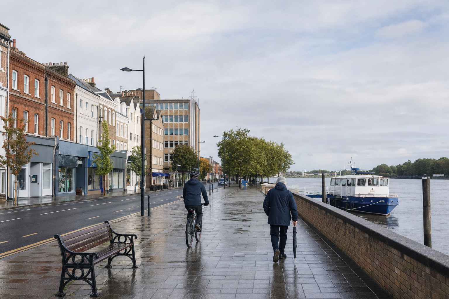 A cyclist and pedestrian with an umbrella on a wet riverside path lined with buildings and trees.