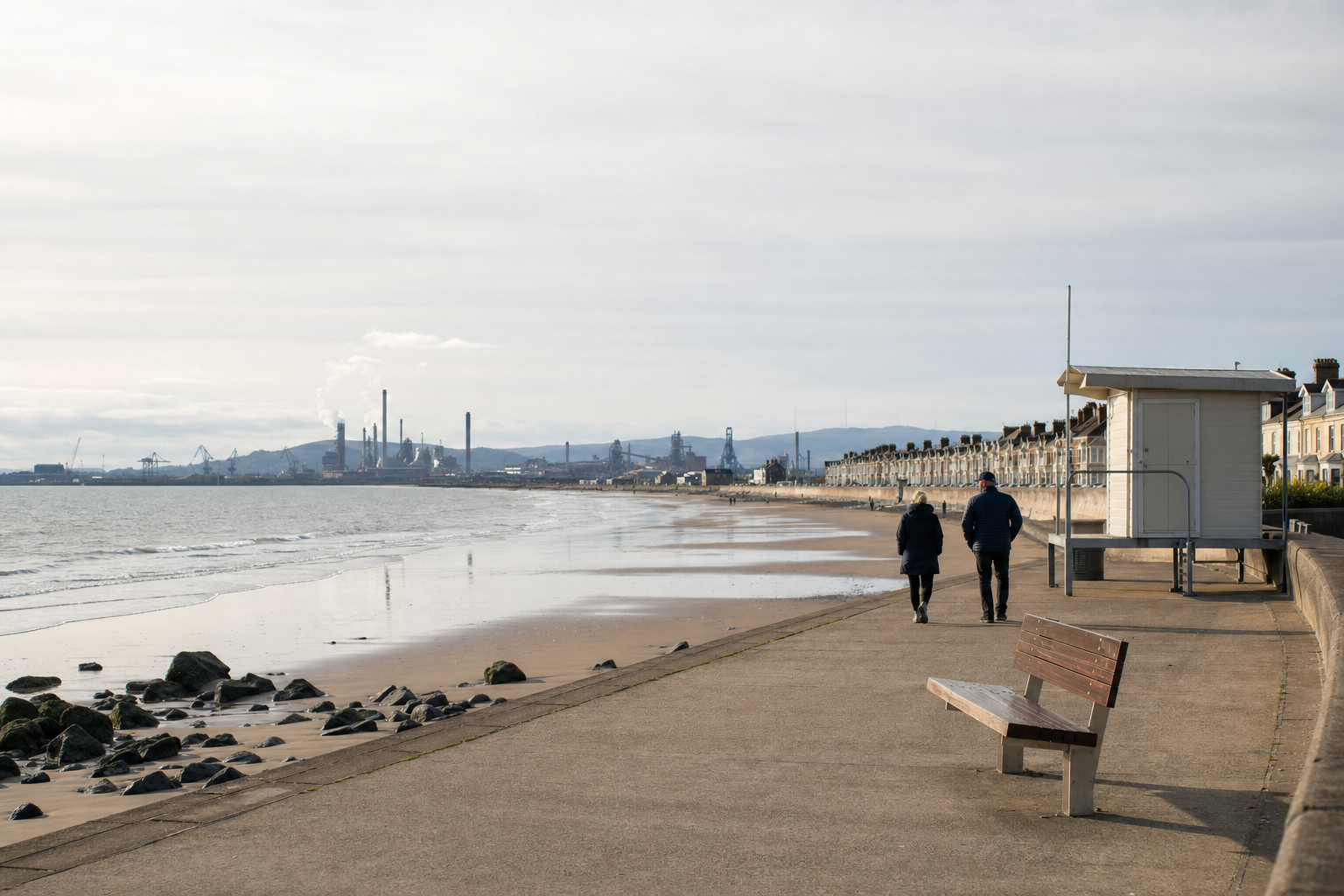Two people walking along a quiet beach promenade with industrial buildings in the background.